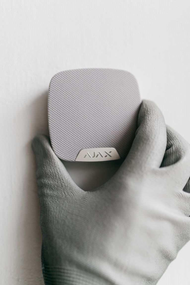 Eine Person mit Handschuhen hält eine Ajax Alarmanlage an die Wand
