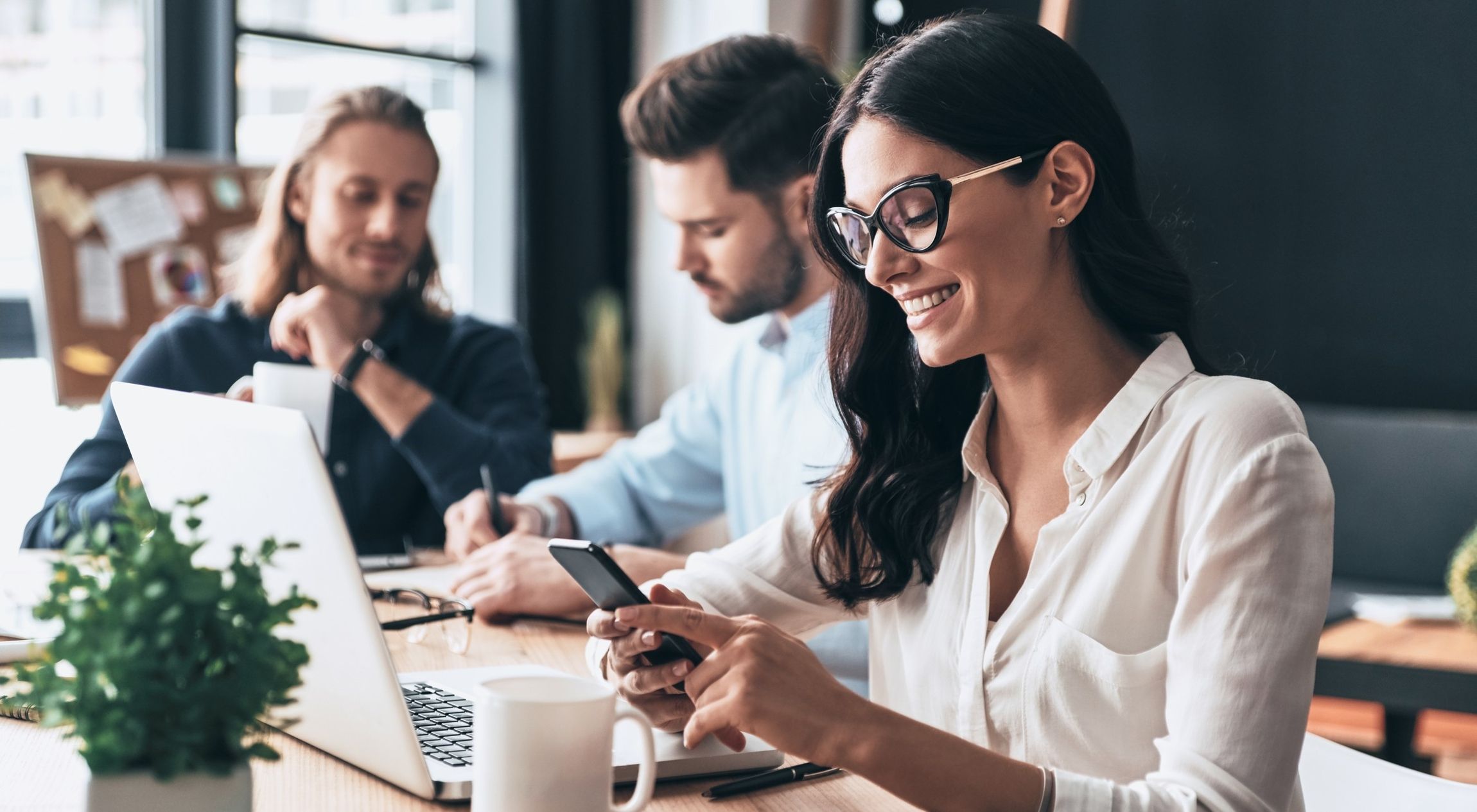 Drei Personen sitzen an einem Tisch mit Laptop, Smartphone und Kaffeetasse in einem Büro mit Pinnwand im Hintergrund